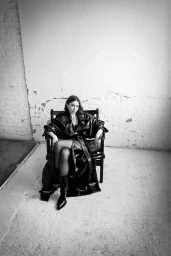 Lily Collins wearing a glossy black leather trench coat and heeled boots, seated on a dark armchair against a distressed white brick wall.