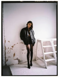 Actress Lily Collins wearing an oversized black leather jacket and tights against a distressed white wall for Elle Spain.