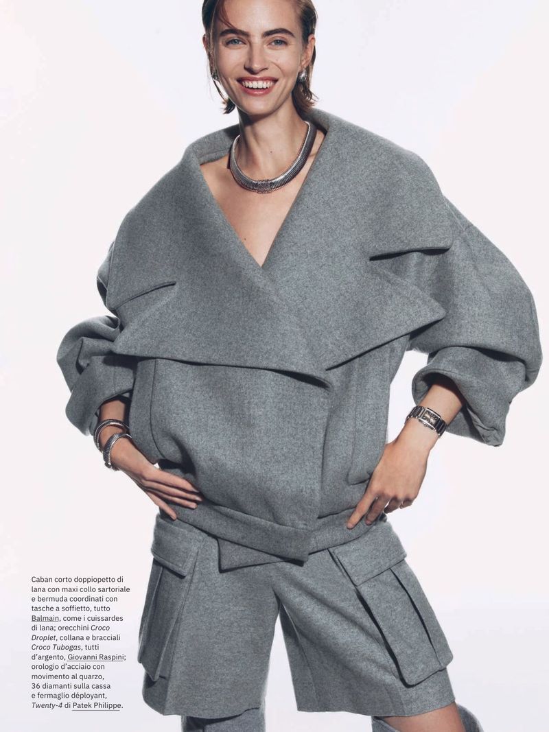 Editorial fashion shot of a smiling adult woman in a grey wool jacket and matching shorts with jewelry against a bright background.