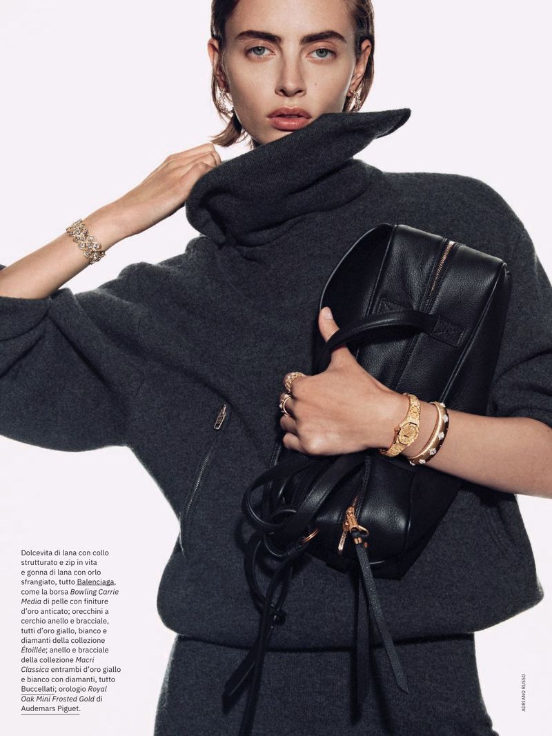A woman poses indoors wearing a gray sweater dress, with gold jewelry, and a black leather bag, shot in medium studio lighting.