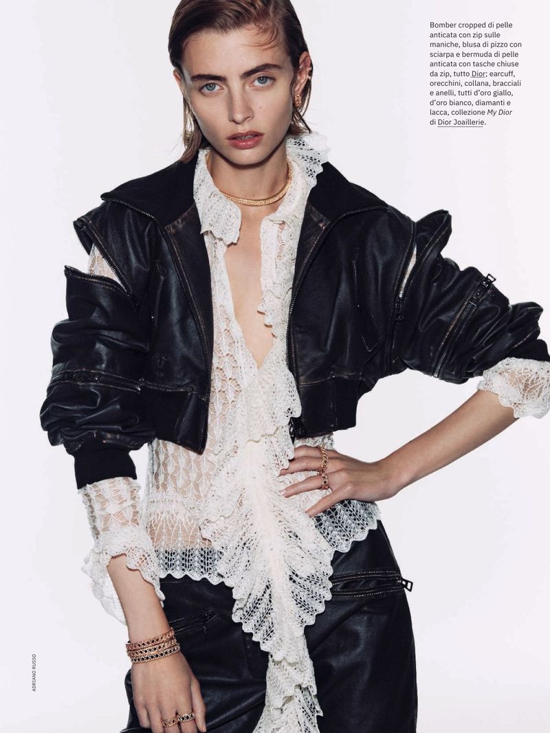 Fashion editorial shot of a young adult woman posing indoors, wearing a black cropped leather jacket, layered with a ruffled blouse.
