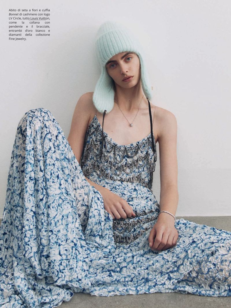 A stylish woman in a floral dress sits against a white wall, complemented by a light blue knit hat and a delicate necklace.