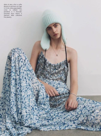A stylish woman in a floral dress sits against a white wall, complemented by a light blue knit hat and a delicate necklace.
