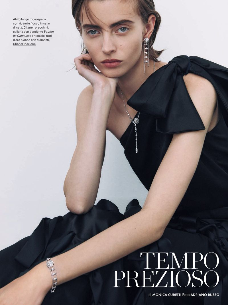 Fashion editorial shot of a brunette woman in a black satin dress and jewelry, posing with hand near her face indoors.