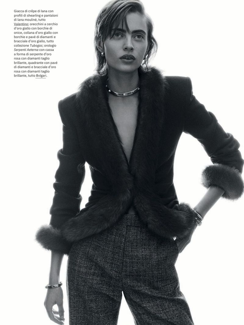 Black and white fashion portrait of a woman in a fur-trimmed jacket and tweed pants with a neutral expression in a photo studio setting.