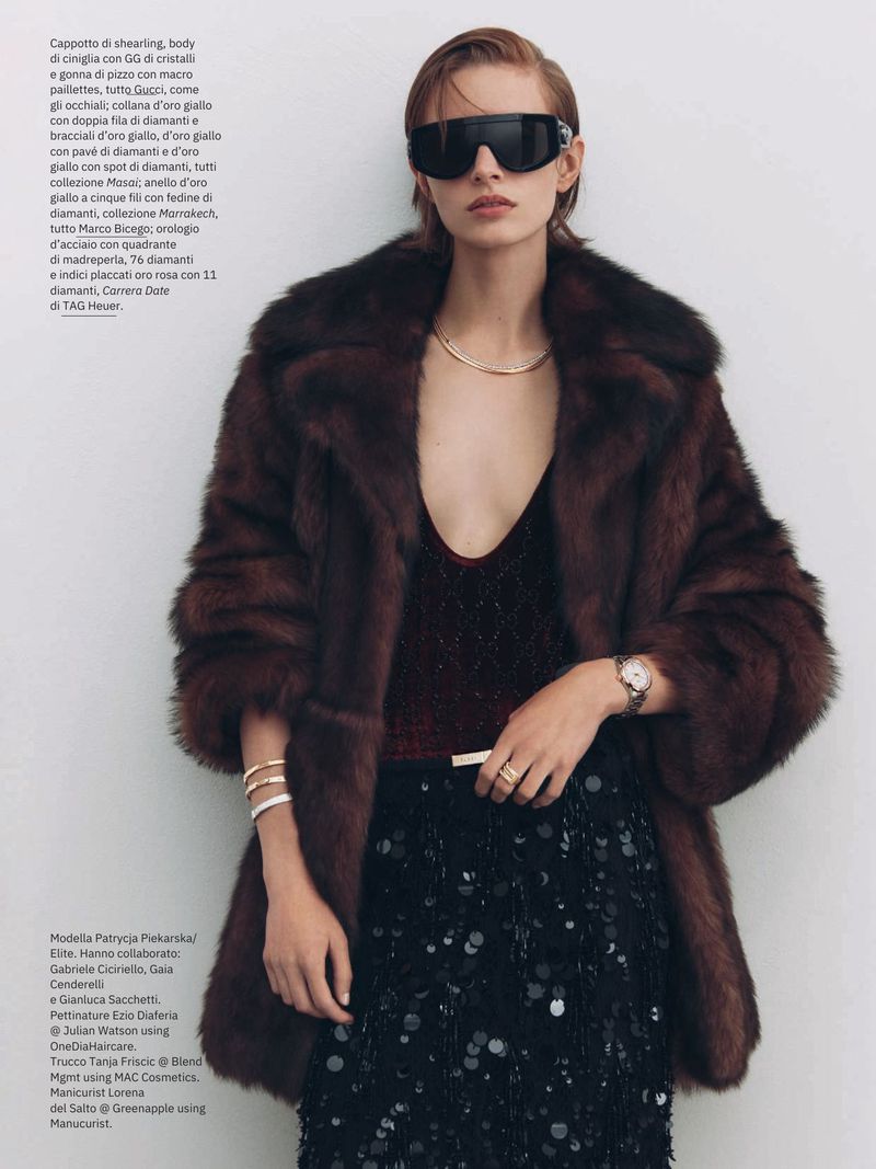 A woman poses in a dark brown fur coat, dark top, and black sequin skirt, accessorized with sunglasses, gold jewelry, and a watch indoors.