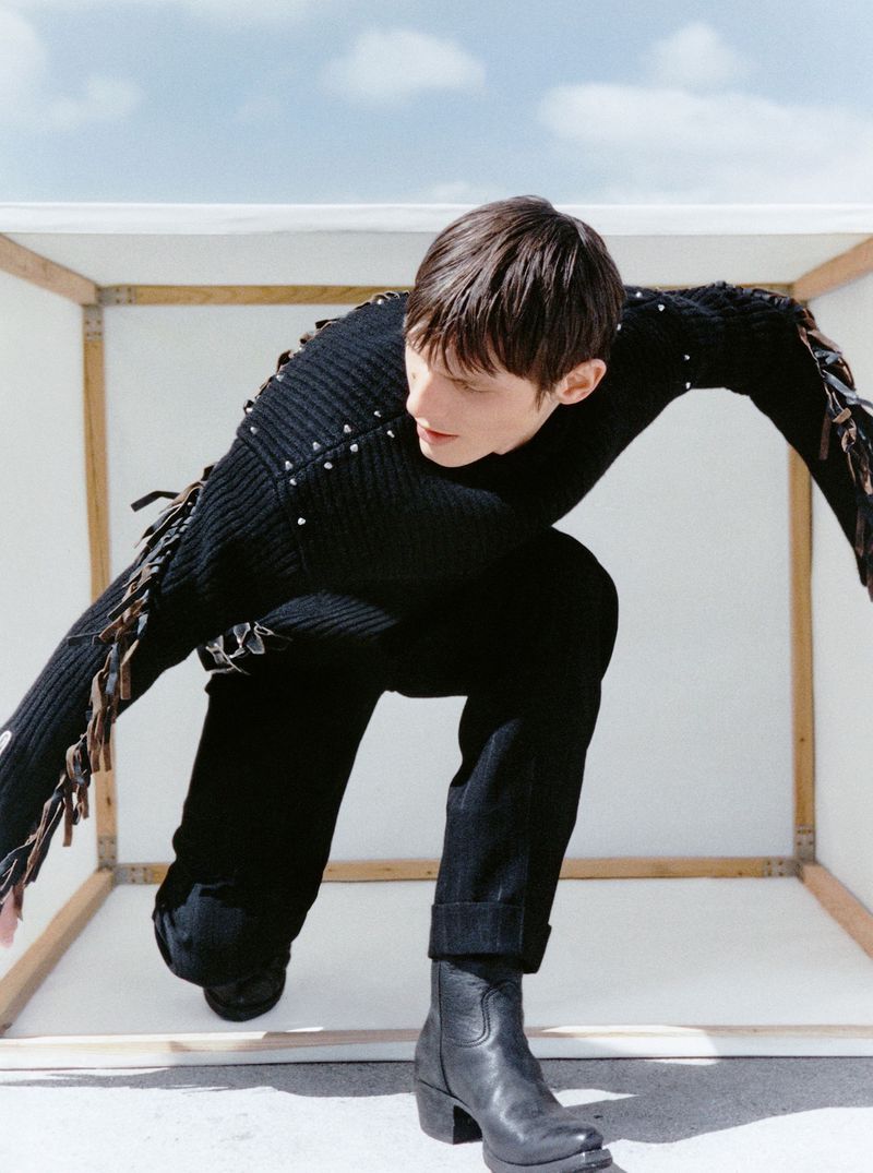 A person in a black sweater and black jeans kneels inside a white box, posing in an outdoor fashion shot with a clear sky overhead.
