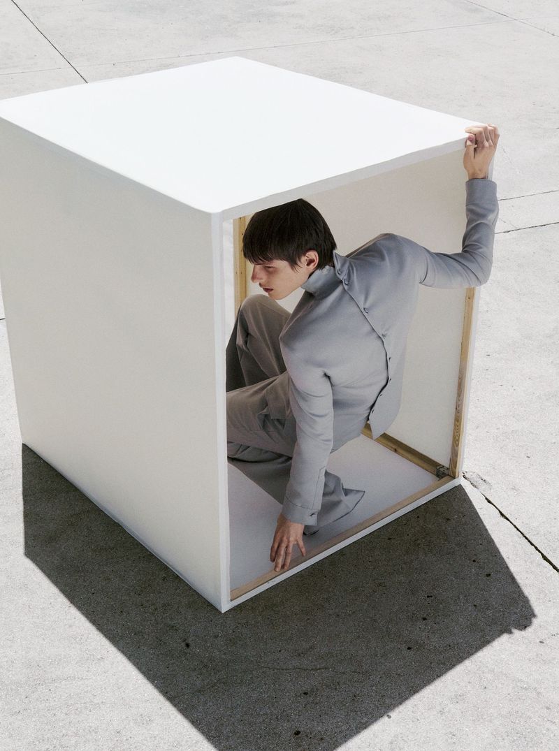 Fashion shot of a young adult in a grey suit, sitting inside a large white cube on a concrete surface in outdoor daylight.