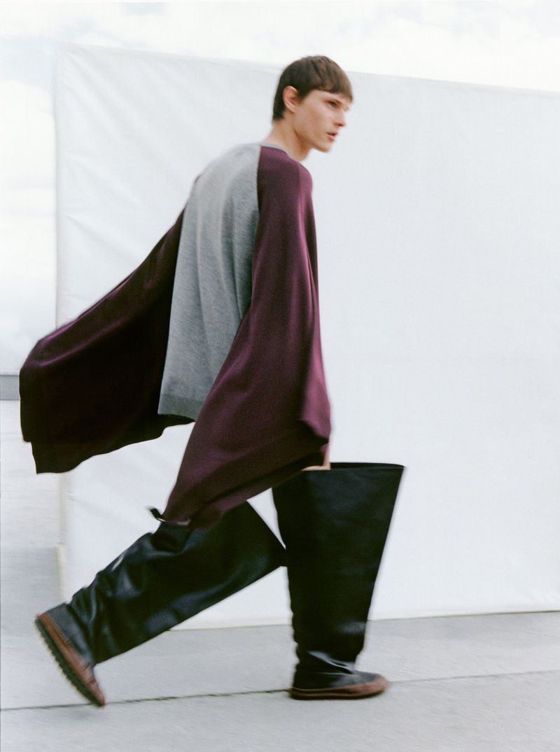 Fashion shot of a male model in motion, wearing a grey top, oversized burgundy sleeves, and black thigh-high boots, set against a white backdrop.