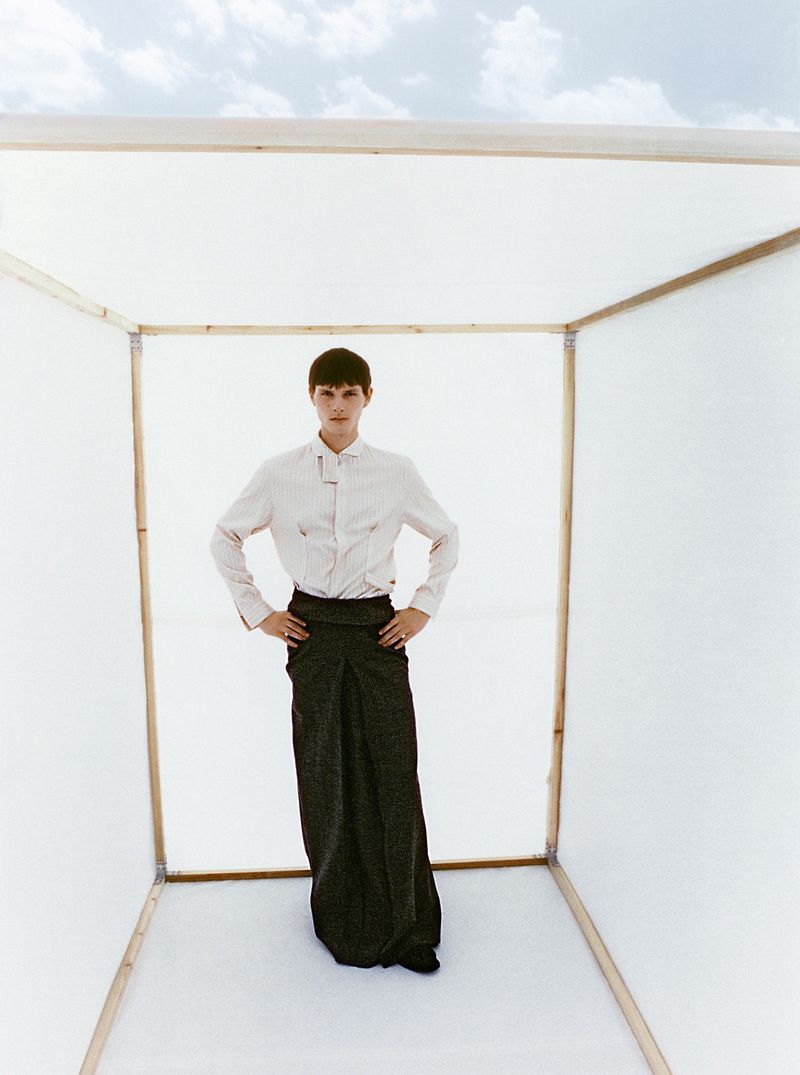 Young man in long wrap skirt and dress shirt poses inside geometric white cube against sky backdrop in high-fashion editorial shot.