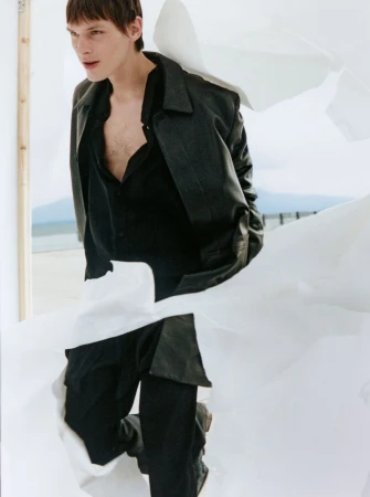 A young adult male model poses in a black jacket and trousers amidst flowing white fabric against a beach landscape backdrop.