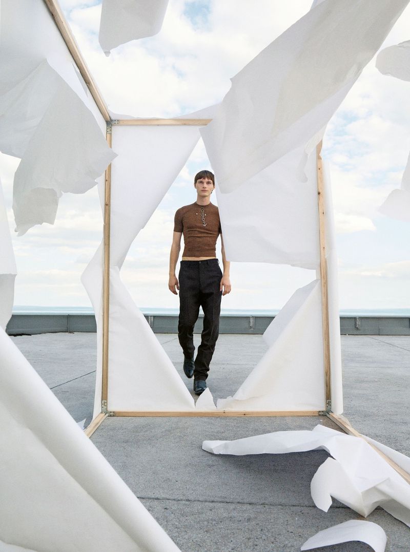 Fashion shot of a young man walking on a rooftop through a makeshift frame with white sheets; sky and ocean visible in background.