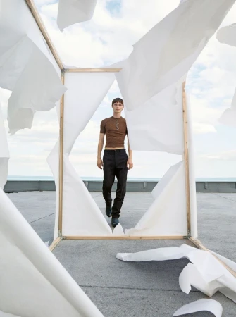 Fashion shot of a young man walking on a rooftop through a makeshift frame with white sheets; sky and ocean visible in background.