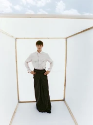 Model Teo Fortin posing with hands on hips wearing a collared shirt and floor-length dark trousers inside a white cubic frame.