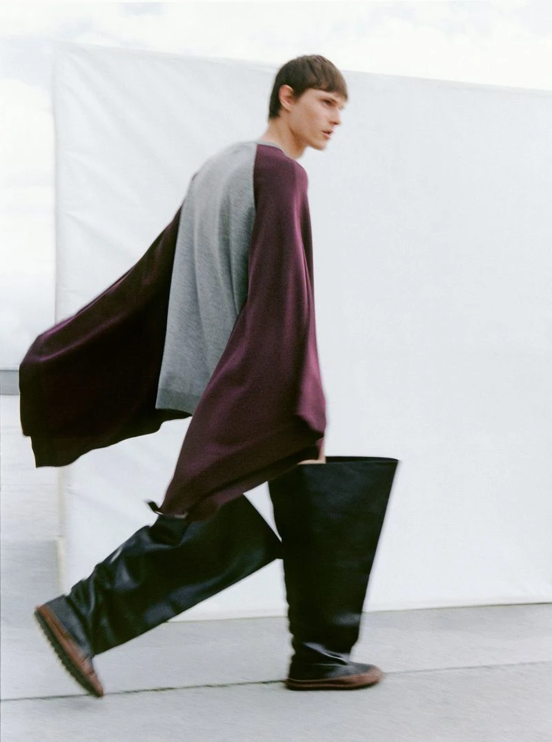 Model Teo Fortin walking in a draped burgundy knit top and wide black leather boots against a white backdrop.