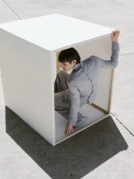 Male model crouching inside an open white cube structure on a concrete surface in an editorial fashion shoot.