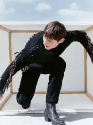 Model in a black textured sweater and dark trousers posing inside a white cube structure outdoors.