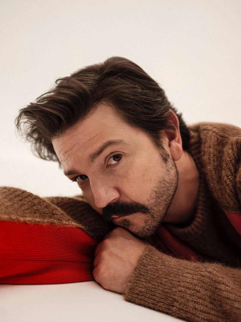 Close-up portrait of a man with a mustache resting his head on a red and brown striped cushion against a plain white background.