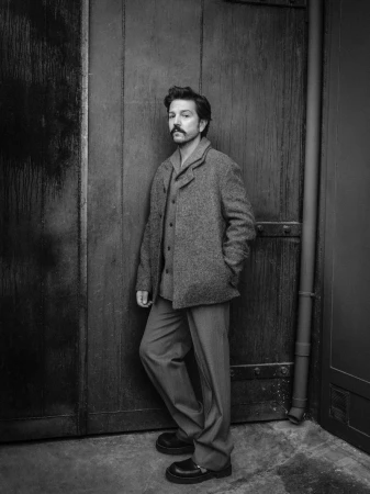 Black and white portrait of actor Diego Luna standing against a dark wooden door wearing a textured wool jacket and trousers.