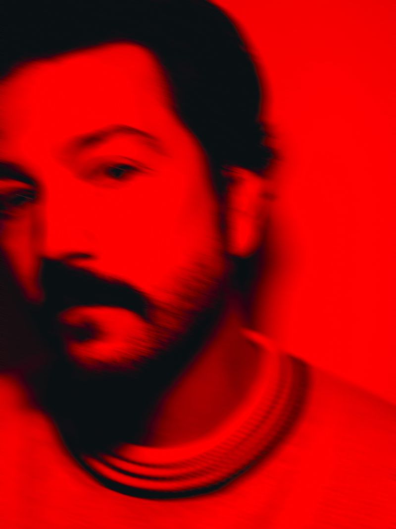 Close-up portrait of a man with a beard and mustache in a red monochromatic color scheme, emphasizing shadow and texture.