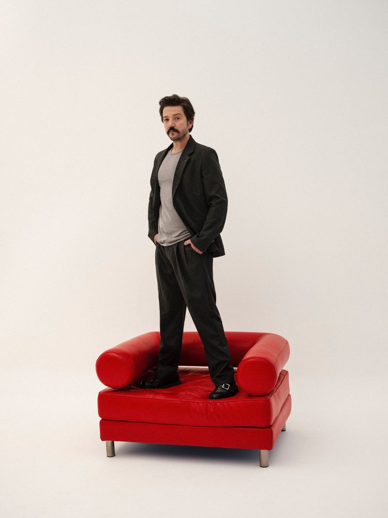 A man poses in a dark grey suit, light grey t-shirt, and black shoes standing on a red chair in a white photo studio setting.