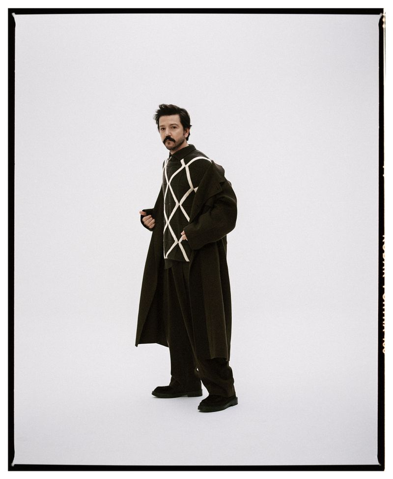 Studio shot of an adult male posing in a dark argyle sweater, matching trousers, and an overcoat against a clean white backdrop.