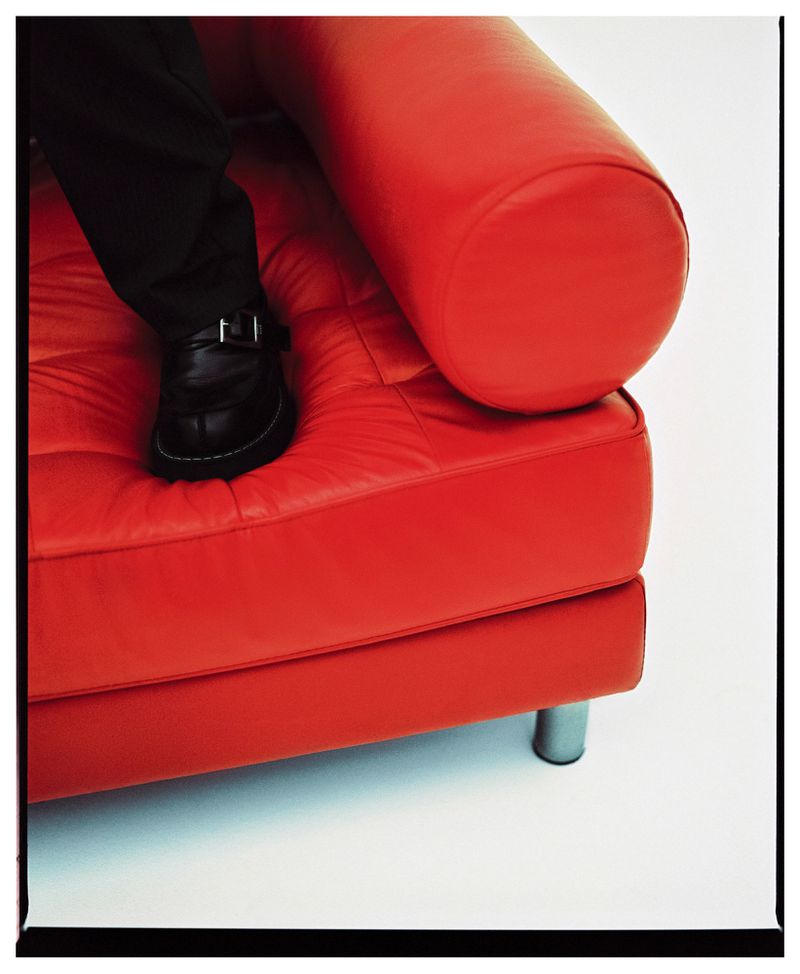 A red leather couch is the focus, with a person's black-trousered leg and black leather shoe stepping onto the cushion in a clean, bright studio.