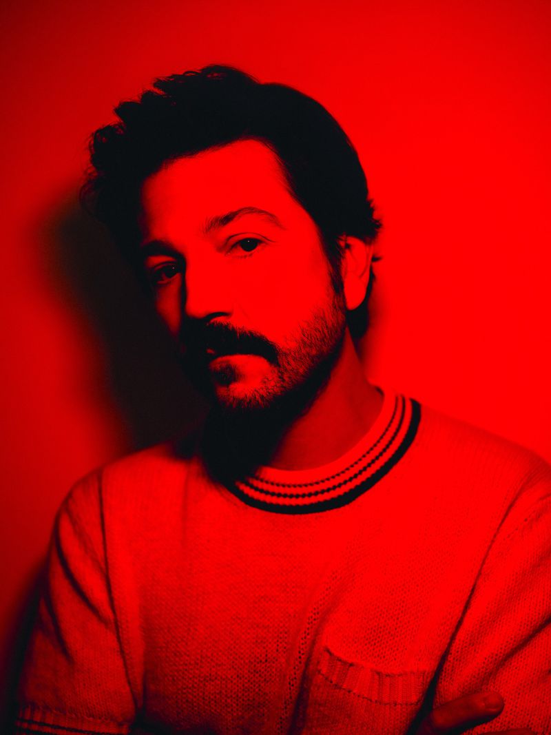 Close-up portrait of an adult male with a mustache, lit with red lighting, wearing a white t-shirt against a red background.
