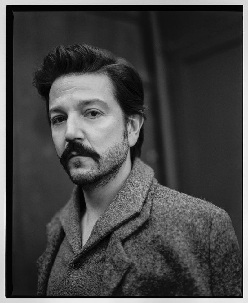 Monochrome portrait of a moustached man with dark hair, wearing a wool coat against a dark background. Studio lighting, close-up.