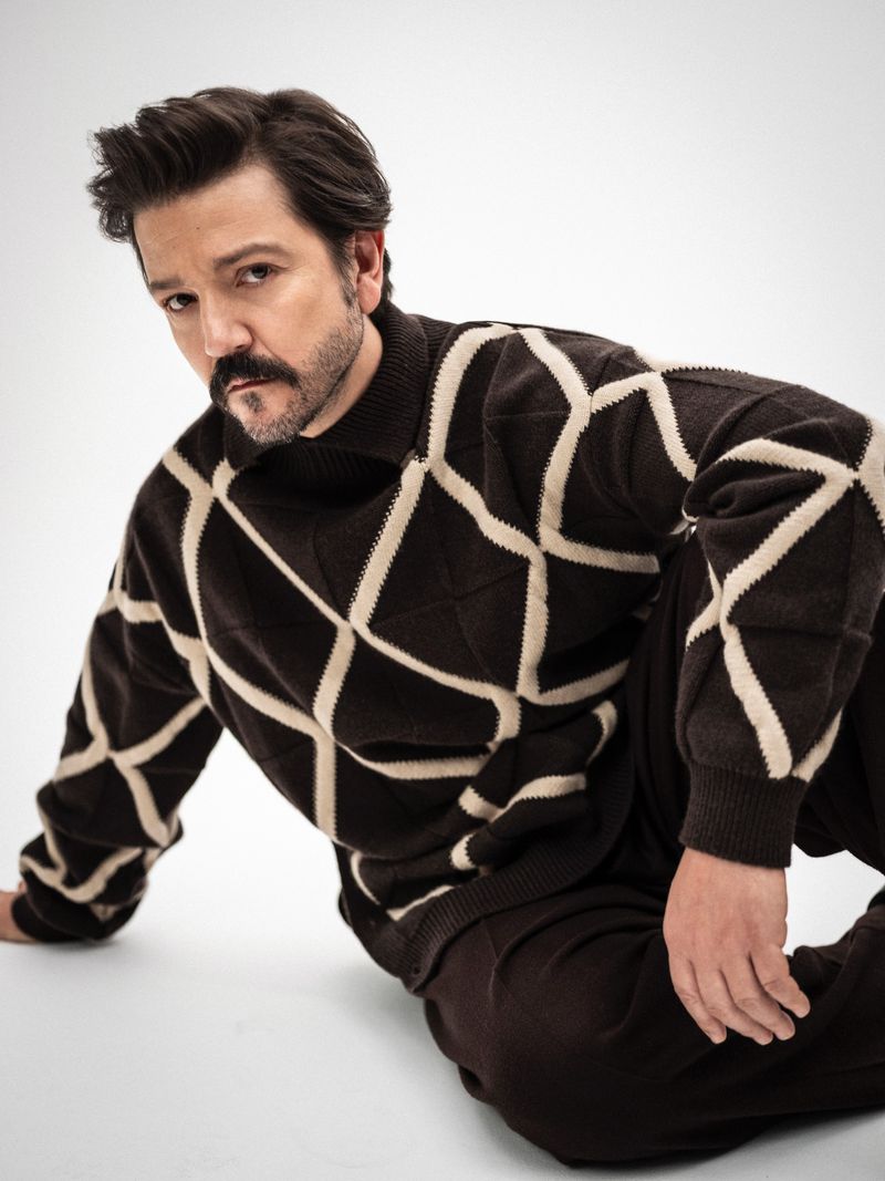 Portrait of adult male wearing a brown and beige patterned knit sweater and dark pants posing in a photo studio against a white backdrop.