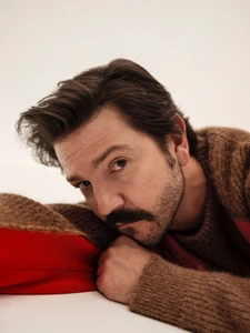 Actor Diego Luna resting his head on his arm while wearing a brown knit sweater with red accents against a white background.