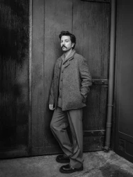 Black and white portrait of actor Diego Luna standing against a dark wooden door wearing a textured wool jacket and trousers.