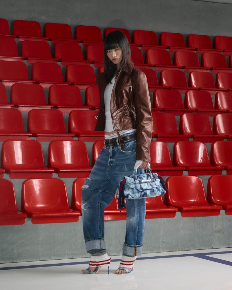 Fashionable woman in a brown jacket and oversized blue jeans poses in front of rows of red stadium seats, holding a blue purse.