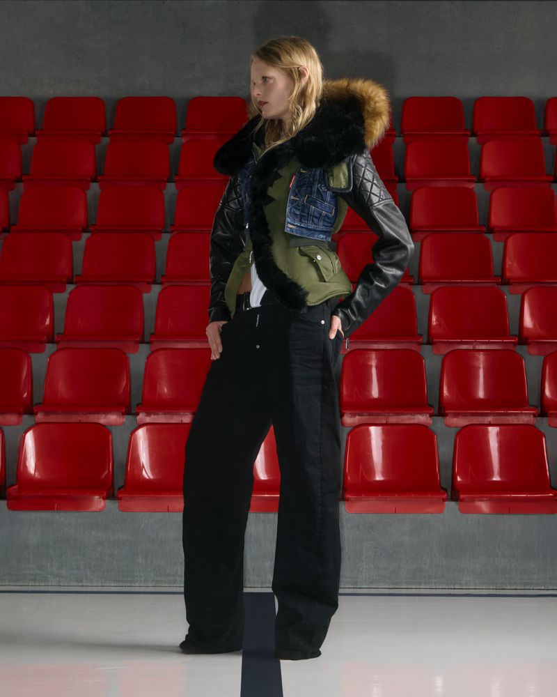 Fashion photo of a blonde model posing indoors wearing a black leather and fur-trimmed coat, black pants, against a red stadium seat backdrop.