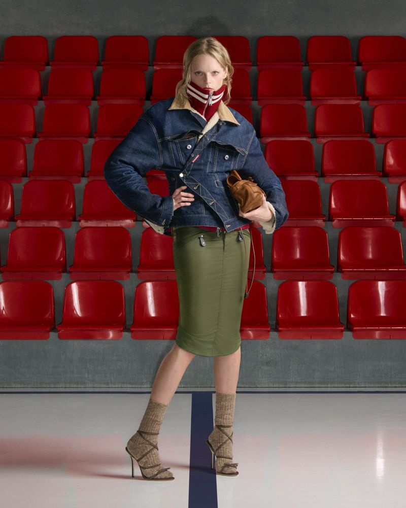 A fashion shot of a woman in a denim jacket, green skirt, and patterned heels, posing in an arena with red stadium seating.