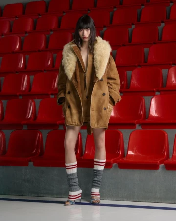 A young woman poses in a brown jacket with a fur collar and grey leg warmers, set against a background of red stadium seats.