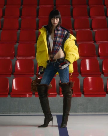 A woman in a yellow jacket and thigh-high boots poses in an indoor stadium with red seats, under even lighting, with a neutral expression.