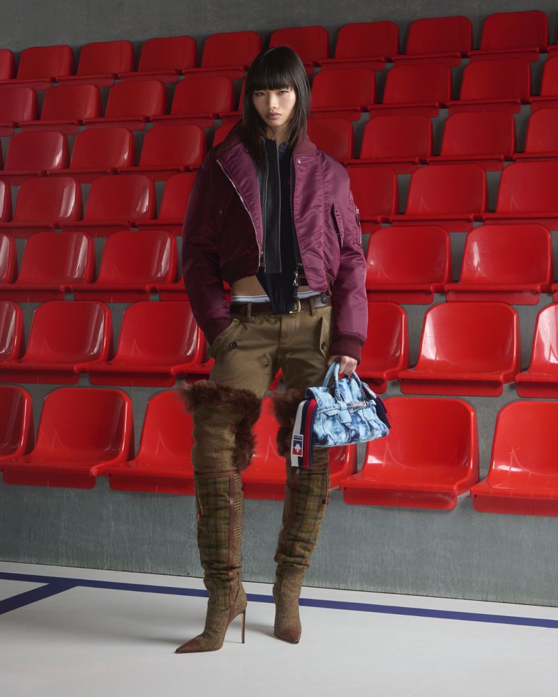 A woman in a burgundy jacket, brown pants, plaid boots, and a denim purse poses in front of red stadium seating for a fashion editorial.