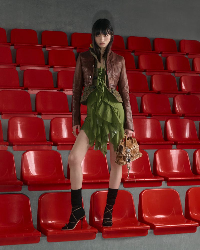 Fashionable Asian woman in green dress, brown jacket and high heels, holding a beaded bag, posing on stadium seating.