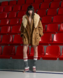 Model Chun Jin wearing a tan shearling coat and striped leg warmers standing before red stadium seats