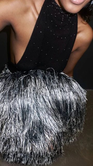 Adult female model poses wearing black and silver dress with tassels. Black backdrop. Shoulder visible, bright light, dark background.