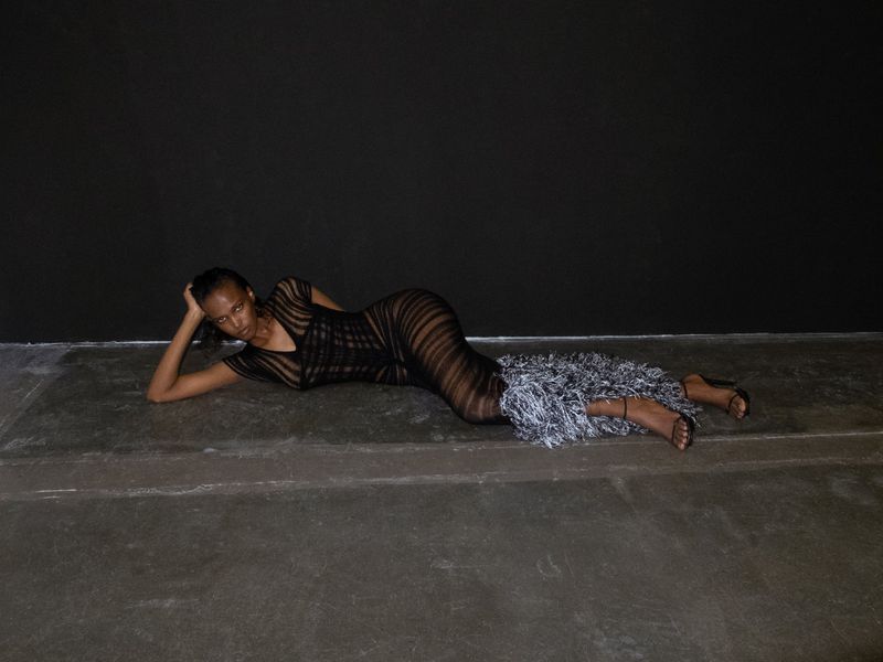 Fashionable woman in a sheer striped dress and strappy heels, lying gracefully on the floor in front of a dark grey background, studio shot.