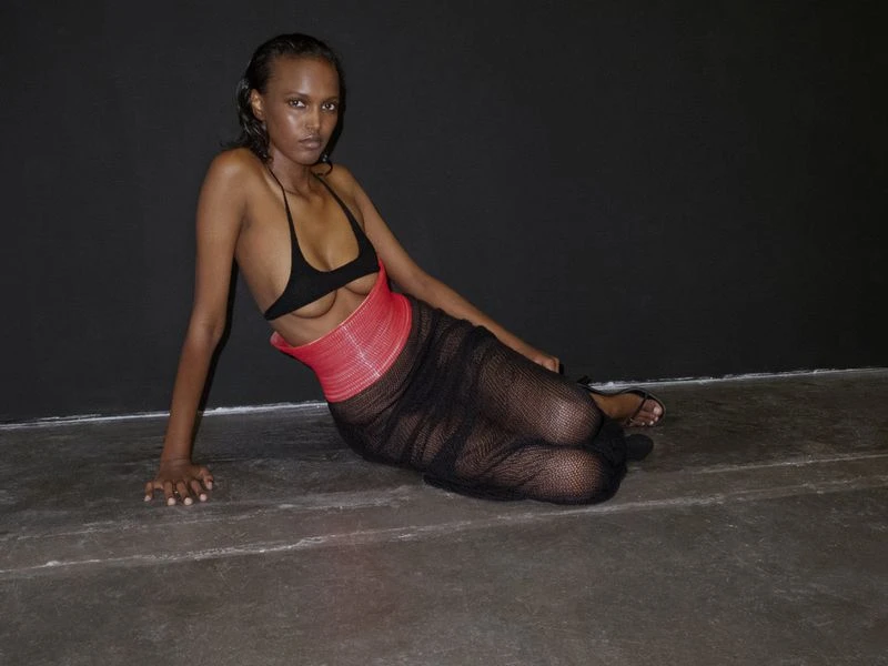 Woman in black bra and mesh dress poses on grey floor against dark grey wall in studio lighting, fashion portrait, low angle.