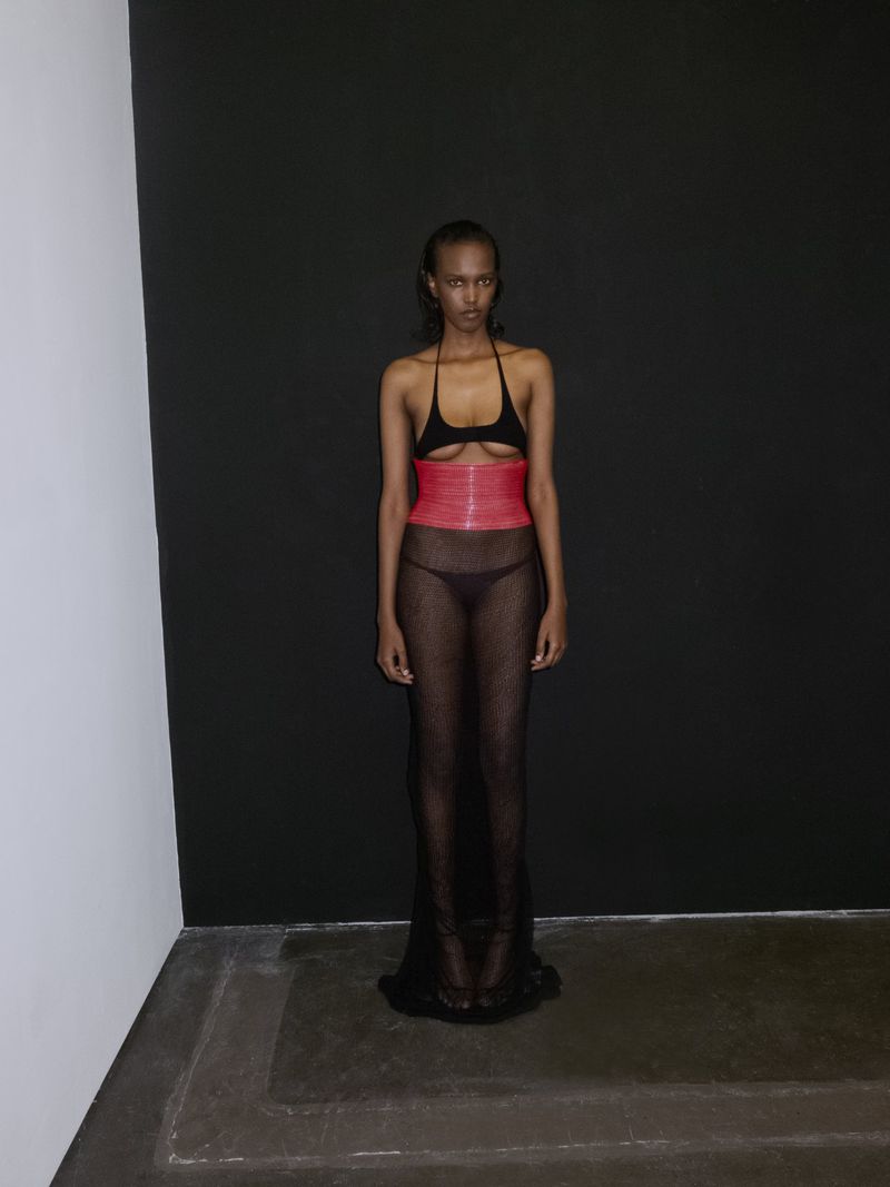 Fashionable young woman poses in a sheer black dress with a red corset in a photo studio setting with black and white walls.