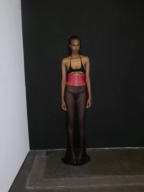 Fashionable young woman poses in a sheer black dress with a red corset in a photo studio setting with black and white walls.