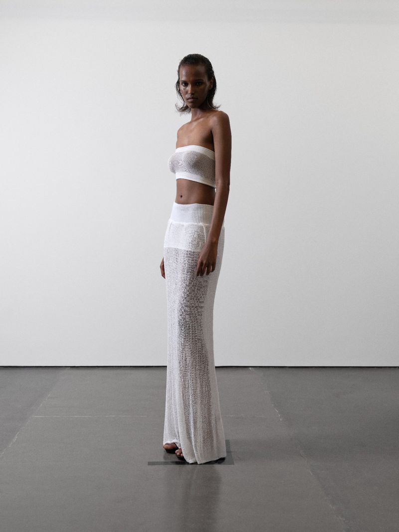 Model Maryamo Musse standing in profile wearing a white sheer knit bandeau top and floor-length maxi skirt against a white studio wall.