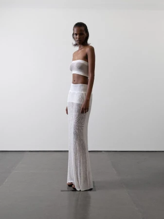 Model Maryamo Musse standing in profile wearing a white sheer knit bandeau top and floor-length maxi skirt against a white studio wall.