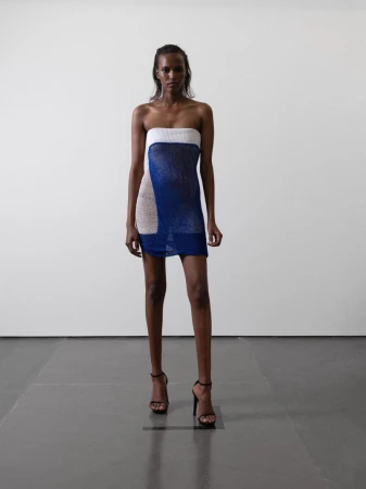 A dark-skinned model poses in a short, strapless knit dress with black high-heeled sandals against a white studio backdrop. Full-length shot.