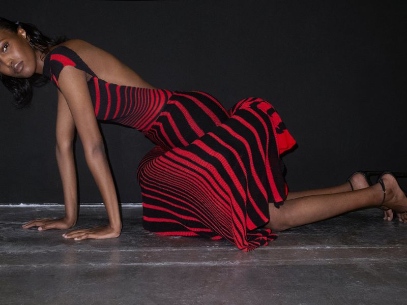 Woman in red and black striped open-back dress poses in a low crouch on a grey surface in front of a black backdrop.