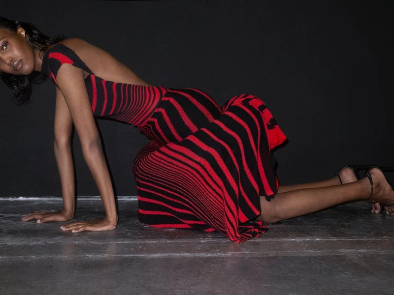 Woman in red and black striped open-back dress poses in a low crouch on a grey surface in front of a black backdrop.